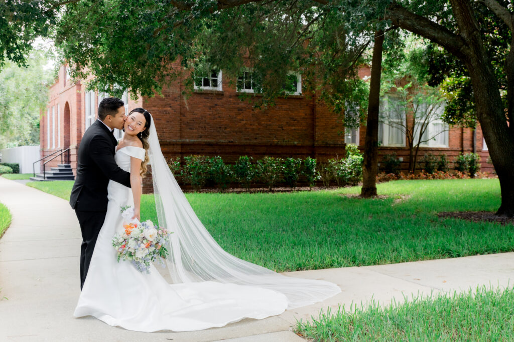 Groom kisses smiling bride on the cheek as she holds a pastel bouquet and wears a long veil and off-the-shoulder gown in front of the red-brick Venue 1902 building on a cloudy wedding day.